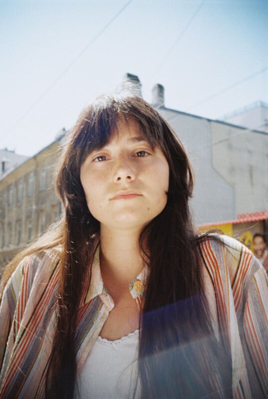 A young woman with long brown hair and bangs, wearing a striped shirt over a white top, looks at the camera outdoors on a sunny day. Buildings are visible in the blurred background.