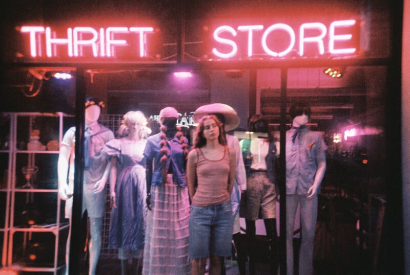 A woman stands in front of a window display with mannequins dressed in various outfits beneath a glowing neon "THRIFT STORE" sign, illuminated by pink and purple lights at night.