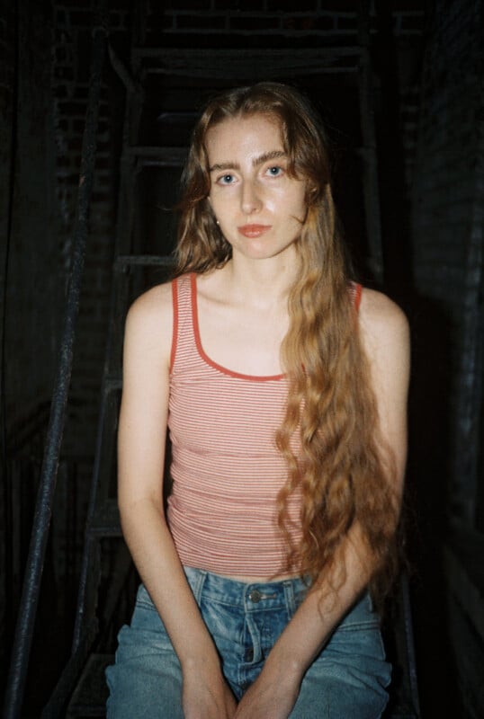 A young woman with long wavy hair sits on metal stairs, wearing a red and white striped tank top and blue jeans, looking at the camera with a neutral expression. The background is dimly lit with brick walls.