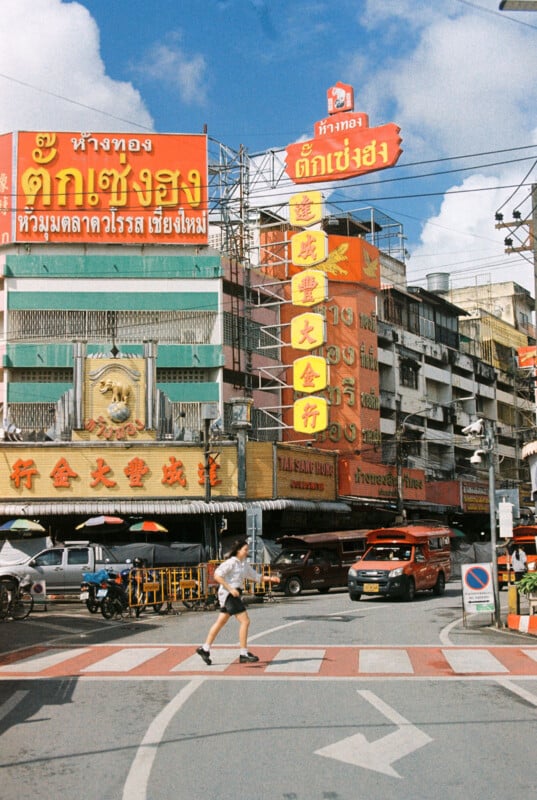 A person in shorts jogs across a crosswalk in a busy city street lined with colorful buildings and signs in Thai and Chinese characters, with cars and tuk-tuks nearby under a bright blue sky.