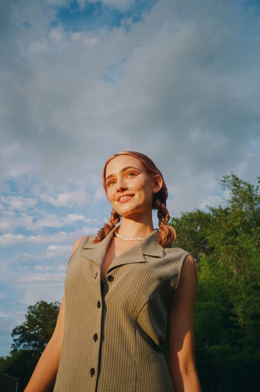 A young woman with braided hair and a sleeveless, button-up dress stands outdoors, smiling under a partly cloudy sky with green trees in the background, bathed in warm sunlight.