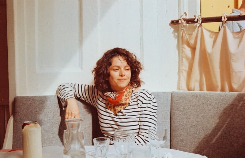 A woman with curly hair and a striped shirt sits on a gray bench in a sunlit room, smiling slightly. There are empty glasses, a candle, and a carafe on the white table in front of her.