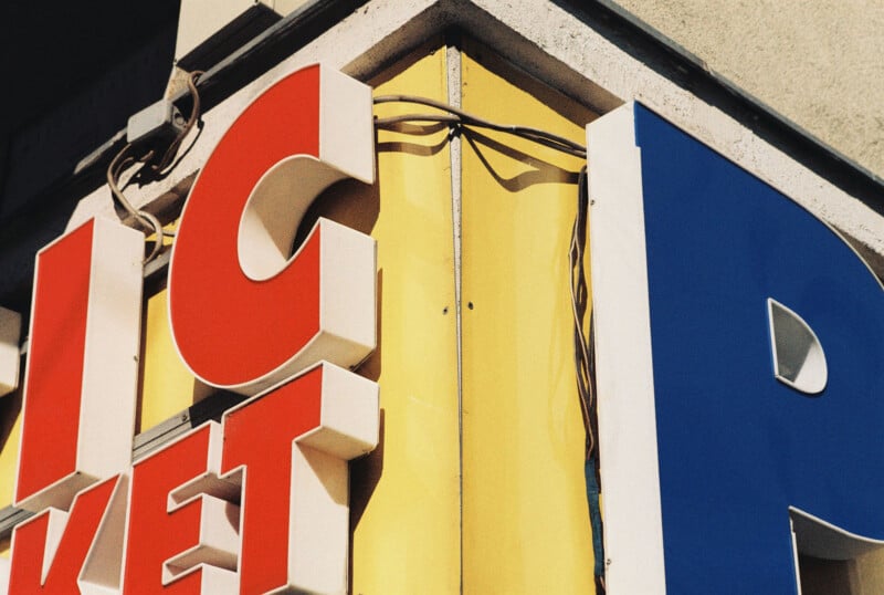 Close-up of a colorful, vintage-style sign on a building corner, showing large red, white, and blue letters, some partially cropped, with visible electrical wiring above a yellow background.