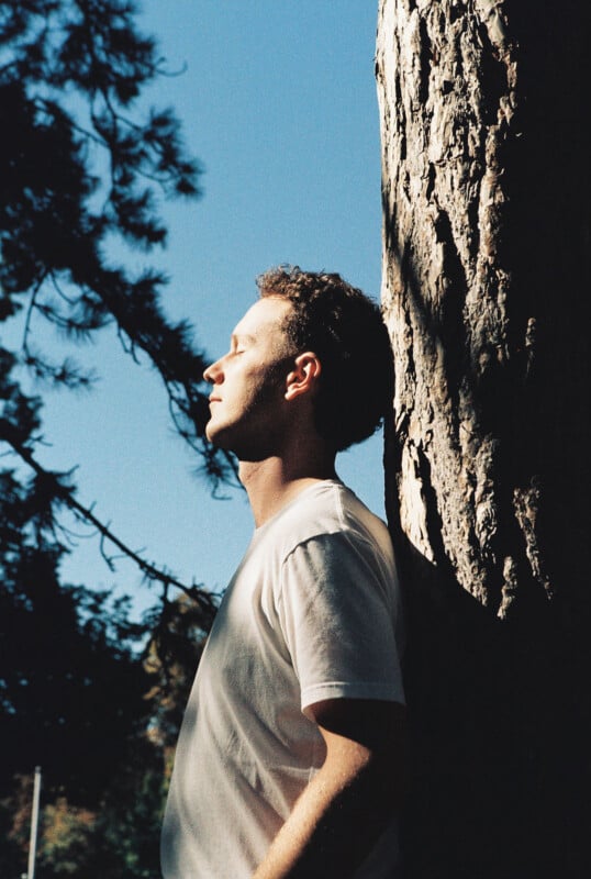 A man in a white t-shirt leans against a tree with his eyes closed, facing the sunlight. The background features blue sky and tree branches, creating a peaceful outdoor atmosphere.