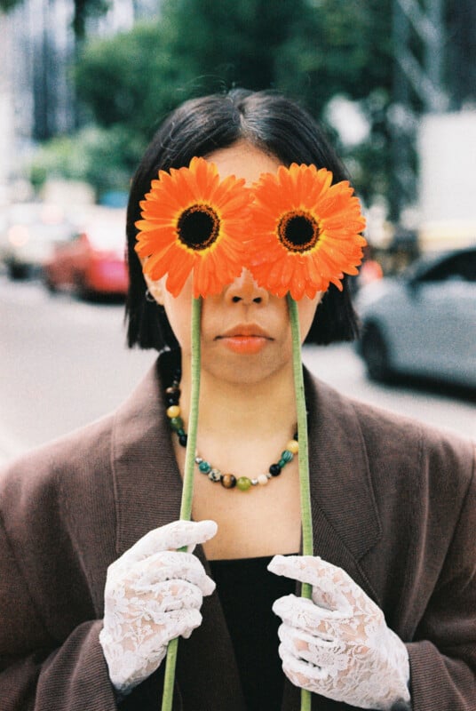 A person wearing a brown coat and lace gloves holds two orange gerbera daisies in front of their eyes, standing on a city street with blurred cars and trees in the background.