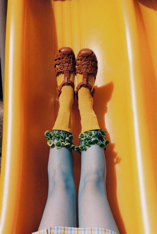 A person slides down a bright yellow slide, wearing blue tights, yellow socks, red sandals, and has curly green kale leaves around their ankles.