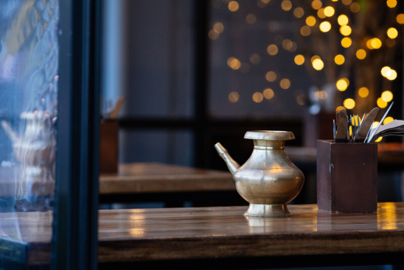 Sobre una mesa de madera del comedor hay una tetera de latón y cerca un recipiente para los cubiertos. Una iluminación cálida y borrosa crea una atmósfera acogedora en el fondo.
