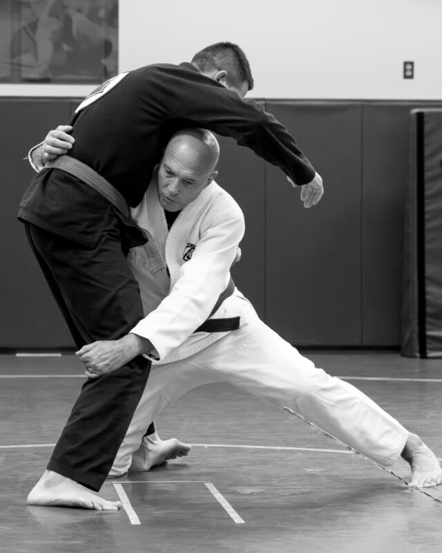 Two men practicing Brazilian Jiu-Jitsu; one in a white gi attempts a takedown by grabbing the other's leg, while the man in a dark gi tries to defend. They are on a mat in a gym. Black and white image.