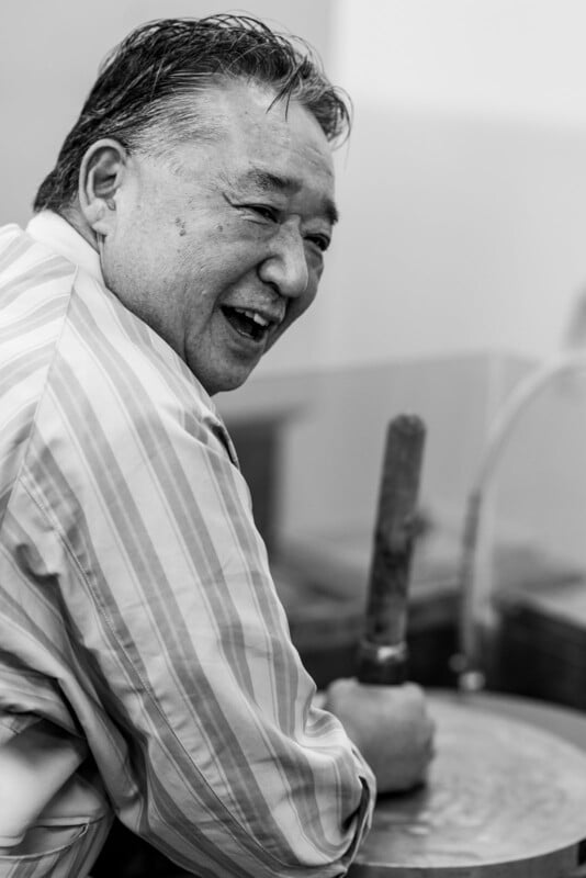 A man in a striped shirt smiles while working with his hands on a circular stone surface, holding a wooden tool. The image is in black and white.