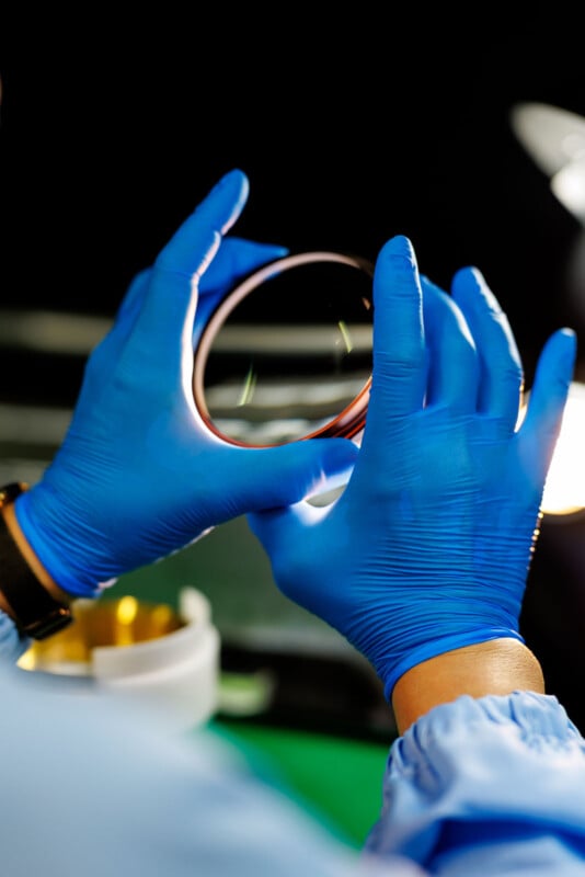 A person wearing blue gloves and a lab coat holds up a petri dish or glass plate, inspecting it closely under bright lighting in a laboratory setting.