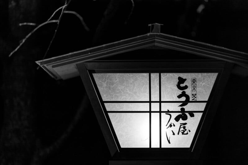 A traditional Japanese lantern with calligraphy on its frosted glass panels glows softly in the dark. Tree branches are faintly visible in the background. The image is in black and white.