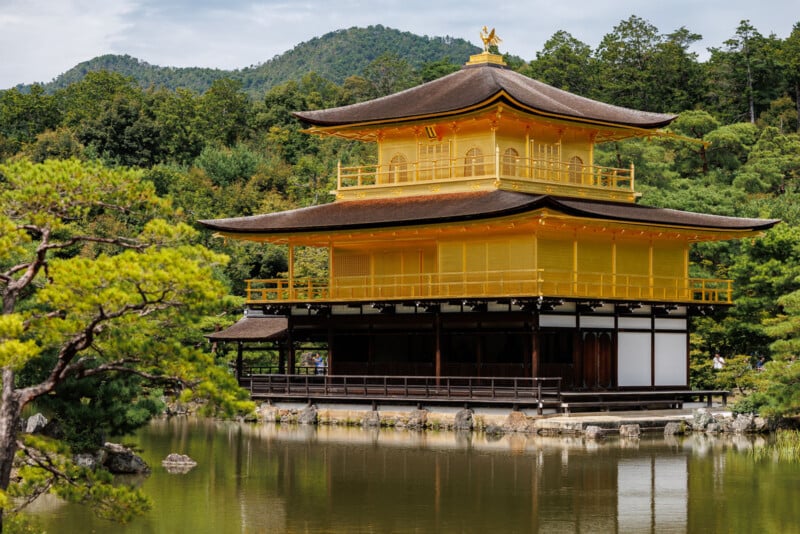 A golden pavilion with two stories stands beside a tranquil pond, surrounded by lush green trees and hills under a partly cloudy sky.