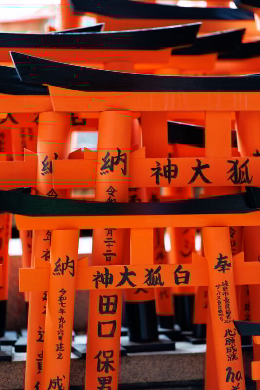 Bright orange torii gates with black Japanese inscriptions are stacked closely together, creating a dense and vibrant pattern often seen at Shinto shrines in Japan.