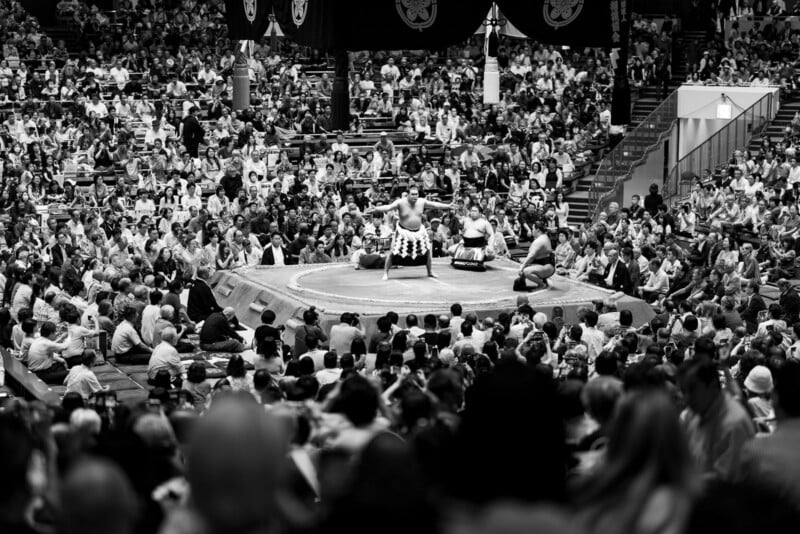 A large crowd watches a sumo wrestling match in an indoor arena. Two sumo wrestlers face each other in a circular ring while a referee stands between them. The scene is in black and white.