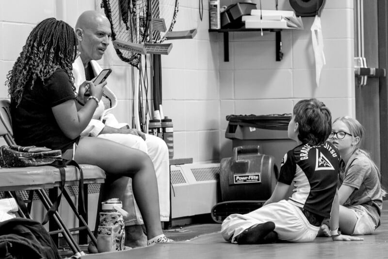 A man in a martial arts uniform talks to two seated women while two children in martial arts attire sit cross-legged on the floor, listening attentively in a gym or training room.