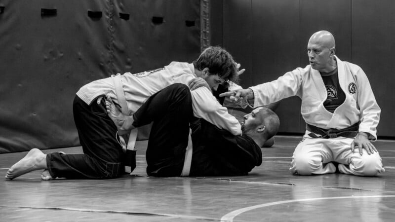 A martial arts instructor in a gi sits on the mat, giving directions to two students practicing grappling techniques during a Brazilian Jiu-Jitsu training session.