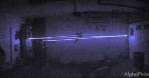 A dark, grainy room with two parallel blue laser beams shining horizontally across the scene. The light creates a glowing effect in the low-light environment.