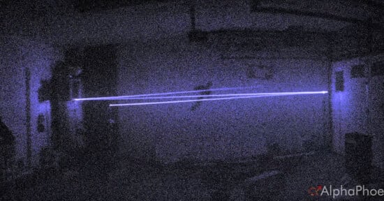 A dark, grainy room with two parallel blue laser beams shining horizontally across the scene. The light creates a glowing effect in the low-light environment.