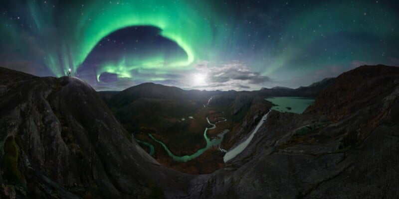 La vibrante aurora boreal verde ilumina el cielo nocturno sobre un paisaje montañoso escarpado con ríos sinuosos, terreno rocoso y lagos remotos bajo un cielo lleno de estrellas.