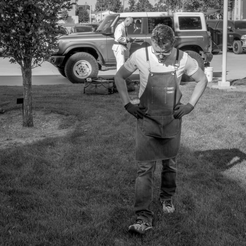 A man wearing an apron and gloves stands on grass, looking down, with a parked Jeep and another person in the background. The scene appears to be outdoors on a sunny day.