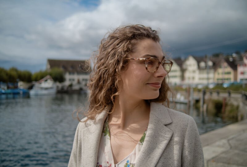 A woman with curly hair and glasses smiles while standing by a waterfront, with boats and buildings in the background under a cloudy sky.
