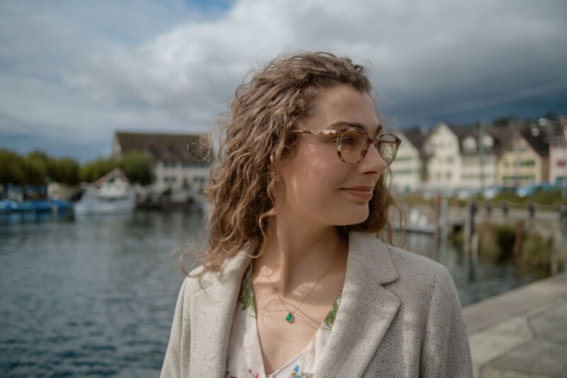 A woman with curly hair and glasses stands near a waterfront, looking to the side and smiling. She wears a light-colored blazer and floral top. Boats and colorful buildings are visible in the background under a cloudy sky.