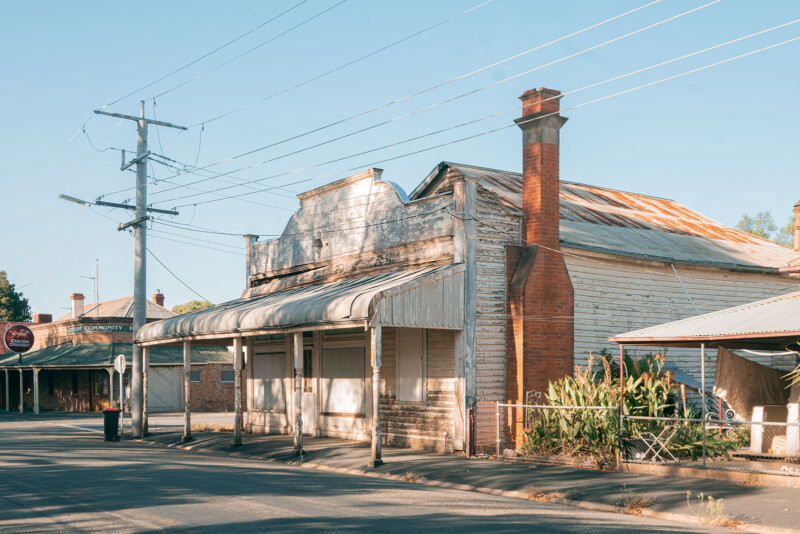 Bajo un cielo azul claro, una calle tranquila y vacía está llena de escaparates de madera viejos y desgastados con pintura descascarada y techos oxidados. En lo alto se ven claramente los cables eléctricos y las chimeneas de ladrillo rojo.