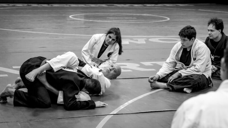 A group of people in martial arts uniforms sit on a mat, attentively watching two individuals demonstrating a grappling technique during a Brazilian Jiu-Jitsu class. The scene is in black and white.