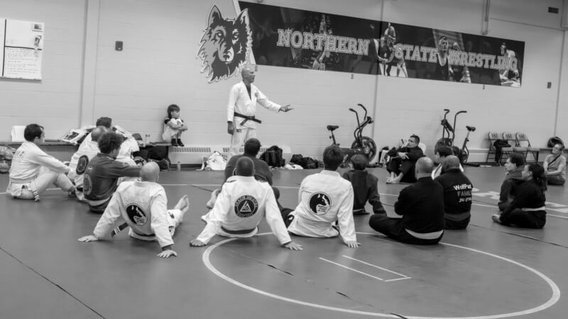 A martial arts instructor stands addressing a group of seated students in gis on a wrestling mat, with a “Northern State Wrestling” sign and exercise bikes visible in the background.