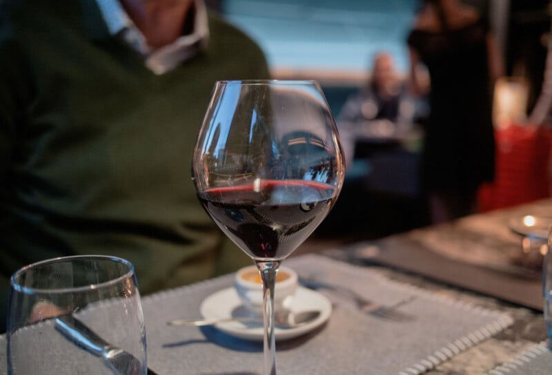 A close-up of a glass of red wine on a dining table, with a blurred background showing a person sitting and another person standing in a restaurant setting. A cup of coffee and a fork are also visible on the table.