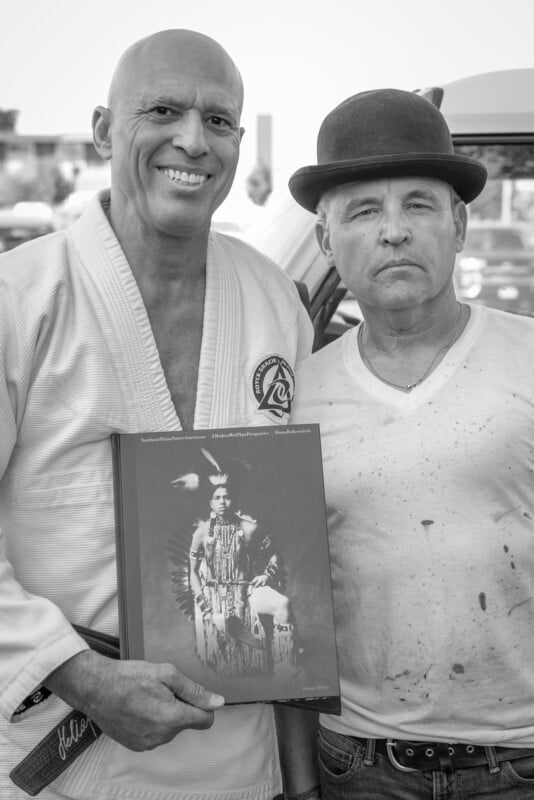 Two men stand side by side; one wears a martial arts gi and smiles while holding a book with a portrait of a Native American on the cover. The other man, wearing a bowler hat and white shirt, looks serious. The photo is in black and white.