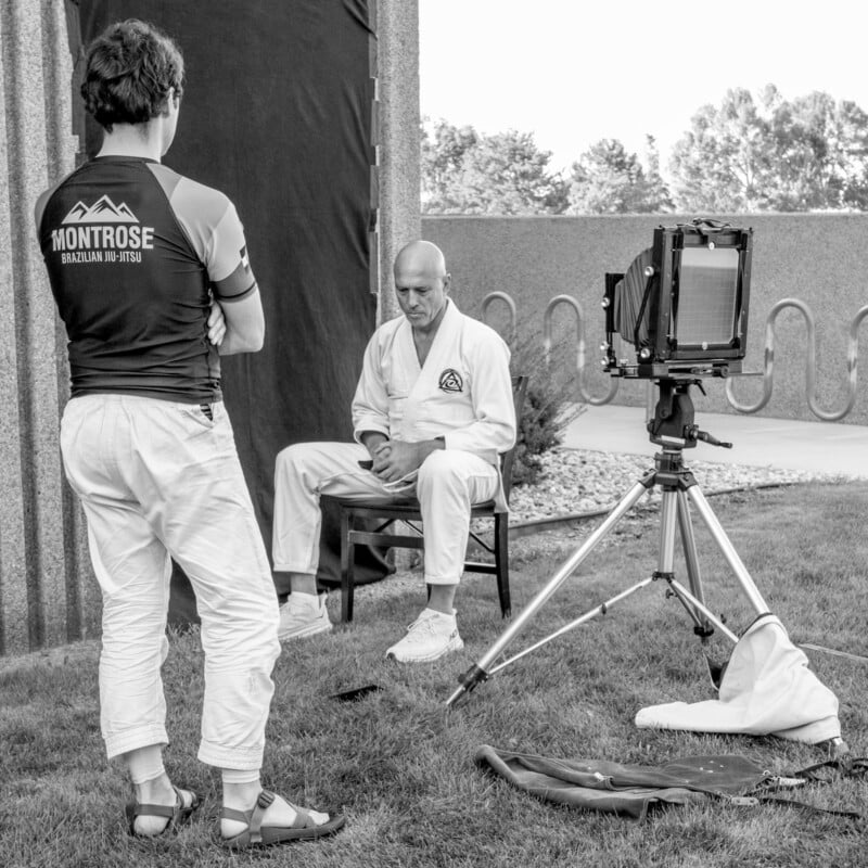 A man in a martial arts gi sits on a chair, posing for a large-format camera outdoors, while another man in a "Montrose Brazilian Jiu Jitsu" shirt stands nearby, watching.