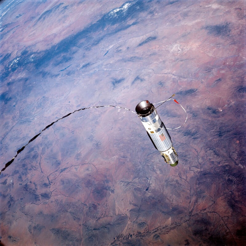 Una nave espacial cilíndrica o un cohete flota sobre la superficie de la Tierra, rastros de materia blanca fluyendo desde sus costados a través del paisaje de terreno marrón y azul que se encuentra debajo.