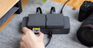 A hand inserts a memory card into a ProGrade card reader with two large memory cards docked, next to a camera and lens on a gray surface.