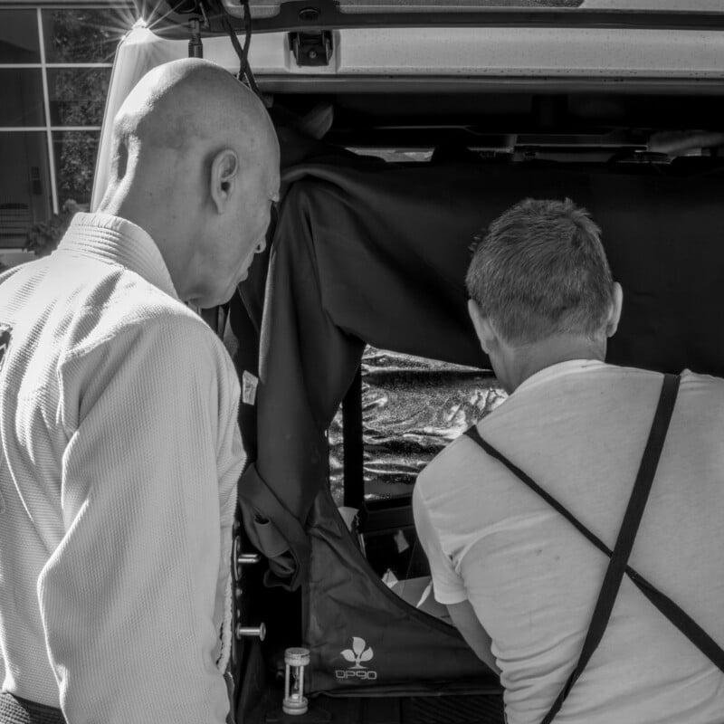 Two people, seen from behind and the side, look into the back of an open vehicle. One wears a white shirt with suspenders; the other is bald and wears a collared shirt. The scene is in black and white.