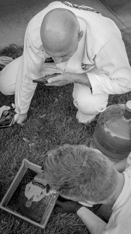 Two people in martial arts uniforms kneel on grass, looking into a tray of water that reflects one person's face. Both hold smartphones, and a large water container is nearby. The image is in black and white.