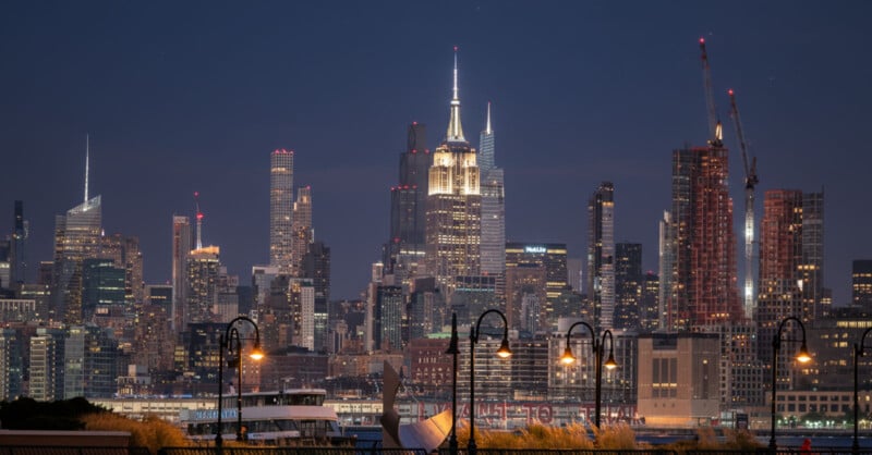 A nighttime view of the New York City skyline with illuminated skyscrapers, including the Empire State Building, and construction cranes, seen behind waterfront street lamps and trees.
