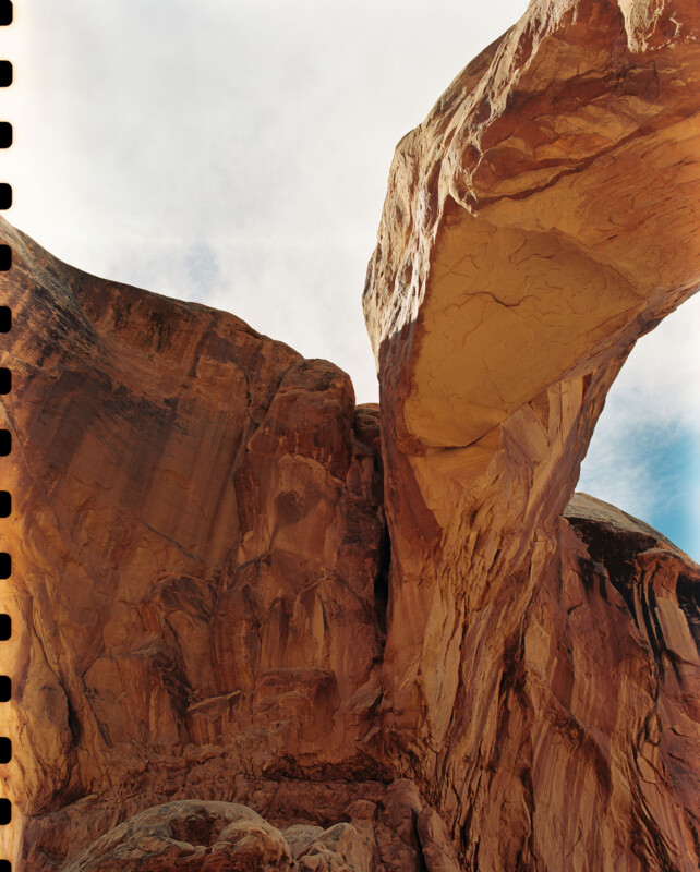 Al mirar el arco de arenisca natural, la vista es espectacular con un cielo parcialmente nublado y cálidas formaciones rocosas de color rojizo. Las perforaciones de la película son visibles a lo largo del borde izquierdo de la imagen.