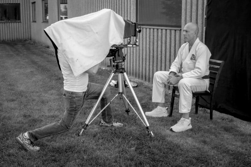 A photographer uses a large-format camera covered by a cloth to take a portrait of a man in a martial arts uniform who sits on a chair outside near a building.