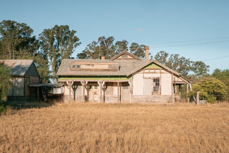 Una casa vieja y desgastada con pintura desconchada y ventanas tapiadas se asienta sobre hierba amarilla seca, rodeada de árboles y cielos azules y despejados.