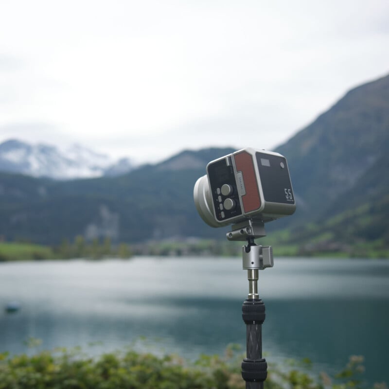 Una cámara antigua montada sobre un trípode enfoca un lago en calma, con colinas verdes y montañas nevadas borrosas en el fondo.
