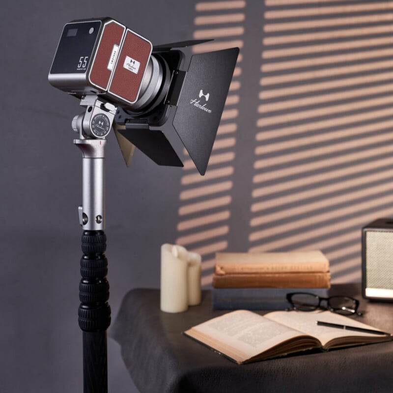 En una habitación cálidamente iluminada, una cámara retro con parasol está montada sobre un trípode. Apilados cerca hay libros, velas, un libro abierto con gafas, un pequeño altavoz sobre la mesa y sombras rayadas en la pared.