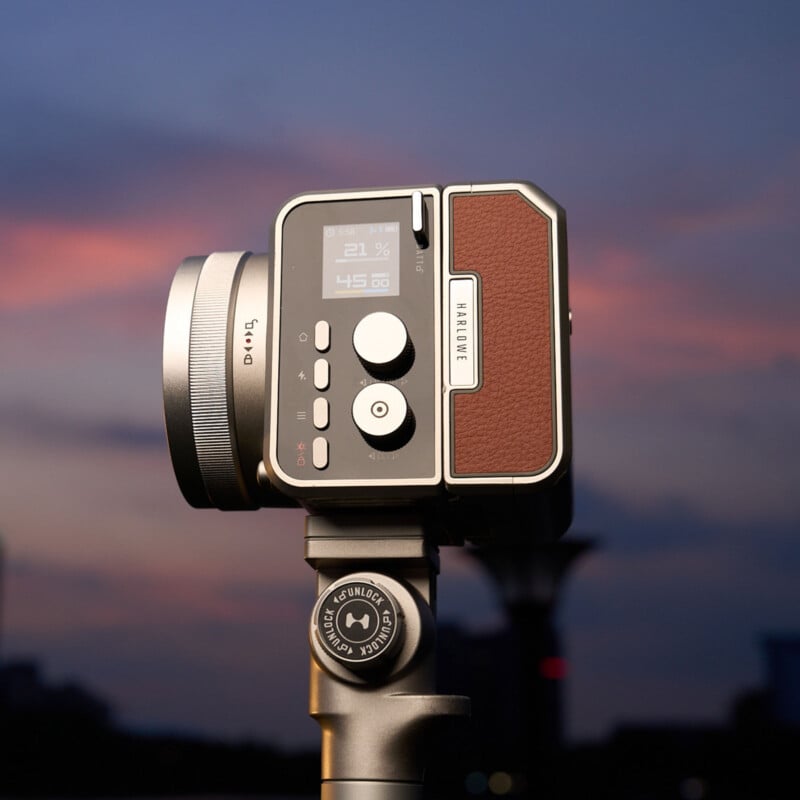 Una cámara digital moderna con un diseño retro con mango de cuero marrón y adornos plateados, montada sobre un trípode contra un colorido cielo al atardecer.