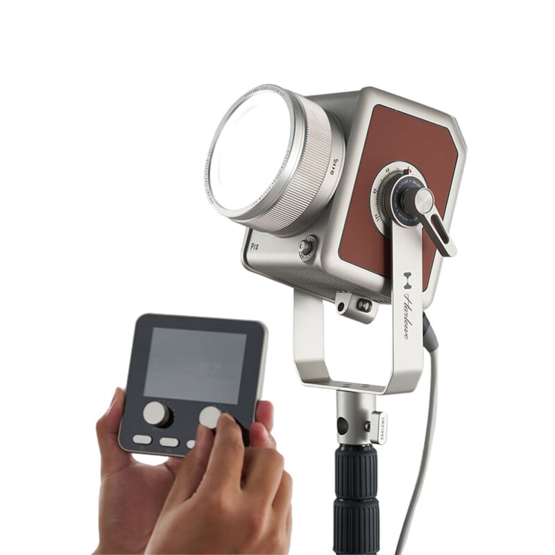 Un hombre está ajustando un controlador digital conectado por cable a una lámpara de estudio cuadrada plateada y marrón de estilo retro montada sobre un trípode sobre un fondo blanco.