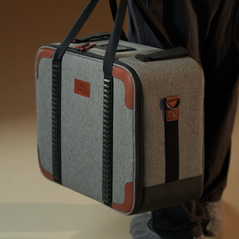 Una persona sosteniendo una maleta de tela gris con correas negras, ribetes de cuero marrón en las esquinas y detalles negros, de pie sobre un fondo neutro.