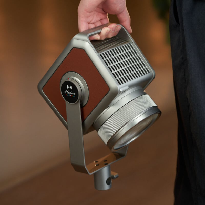 Una persona sostiene un moderno proyector portátil en forma de cubo con mango de metal, paneles laterales marrones, diseño ventilado y una gran lente frontal. El fondo está desenfocado.