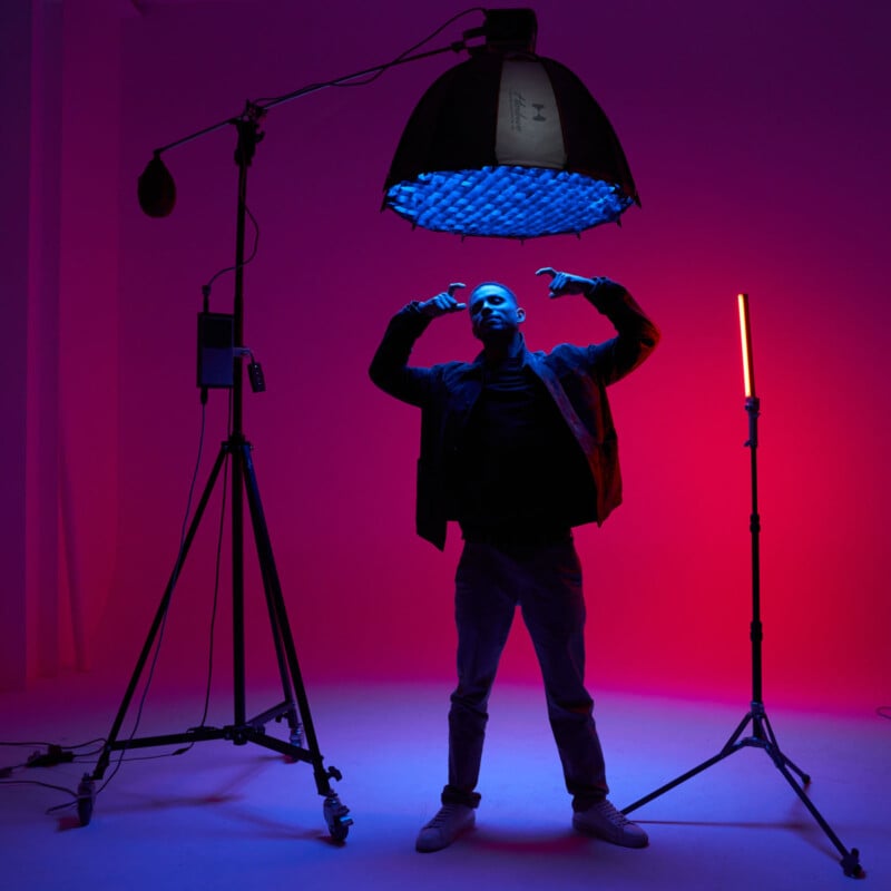 En el estudio, una persona se encuentra bajo una gran lámpara cenital que brilla en vibrantes rosas y azules. Posan con los brazos en alto. Alrededor del perímetro se instalan luminarias y luces LED verticales.