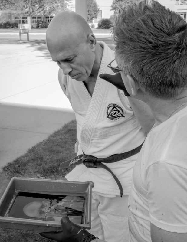 Two men stand outdoors. One, wearing a martial arts gi, looks down at a tray of water held by the other man. The tray reflects the face of the man in the gi. The photo is in black and white.