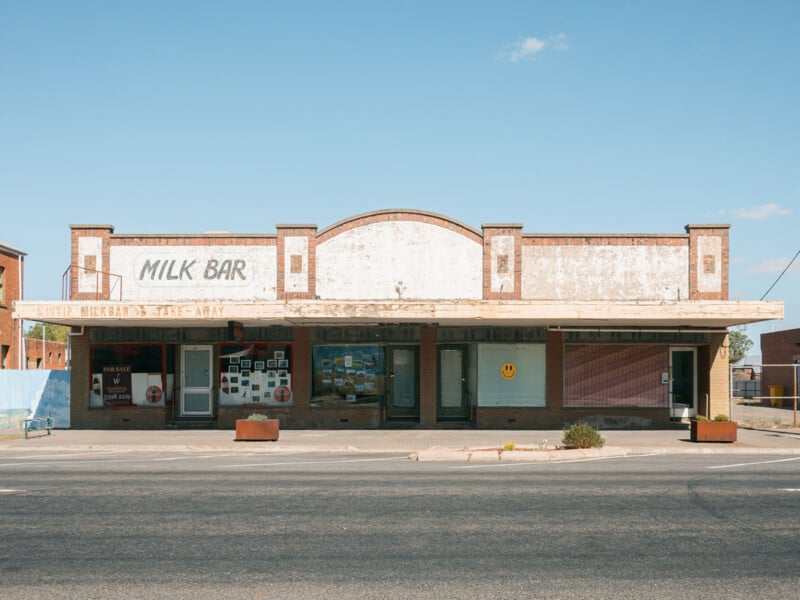 Un viejo edificio desgastado y descolorido "barra de leche" Señalización, un gran ventanal frontal con fotografías, un cartel con una cara sonriente y un amplio alero se alzan sobre una calle vacía bajo un cielo azul claro.