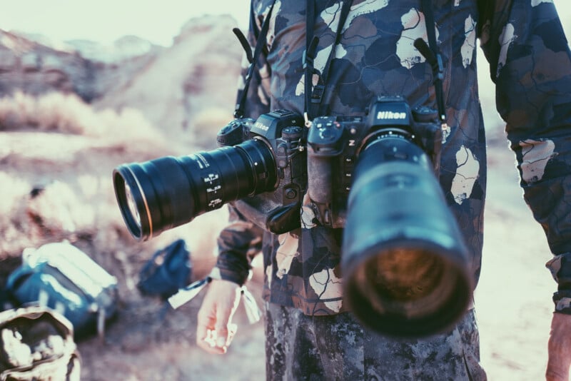 A person wearing camouflage clothing stands outdoors with two Nikon cameras hanging from their neck, one with a wide-angle lens and the other with a long telephoto lens. The background is a rocky, sunlit landscape.